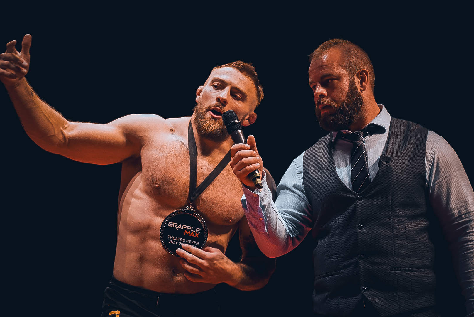 GrappleMax champion on the mic holding the official medal, theatre crowd behind